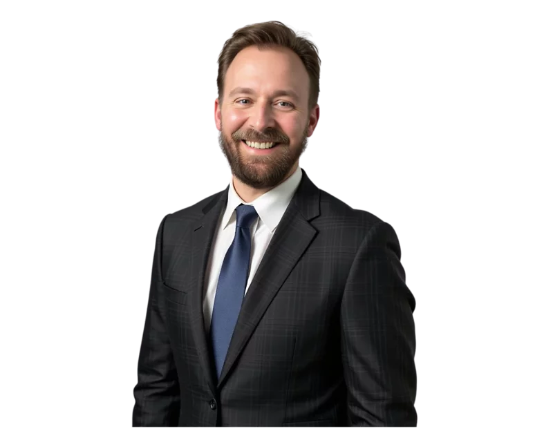 A professionally dressed man with a friendly smile, wearing a dark plaid suit, white shirt, and navy tie, stands against a transparent background. His well-groomed beard and confident posture suggest a corporate or business setting.