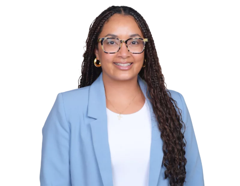 Professional headshot of Ariel Valerio-Meek smiling, wearing glasses, gold hoop earrings, a white top, and a light blue blazer, with long braids draped over one shoulder against a softly blurred light background.