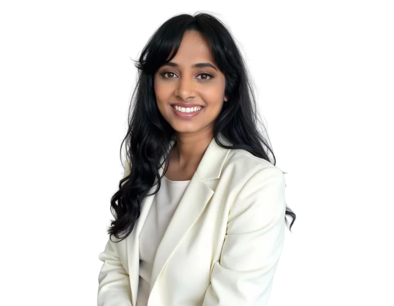 Deepa Kollipara smiling while seated, wearing a cream blazer and blouse, with long wavy black hair against a transparent background.