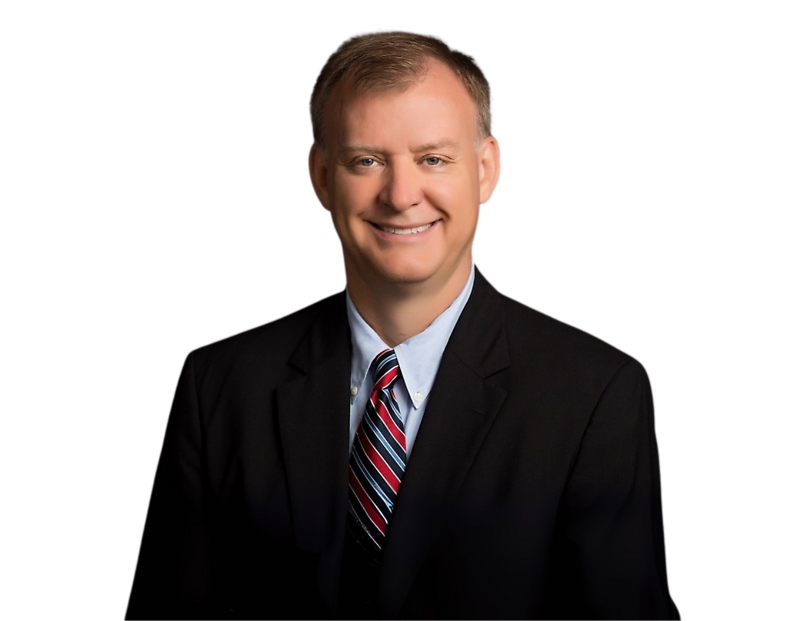 Man with short light brown hair smiling at the camera, wearing a black suit jacket, light blue shirt, and a red, white, and blue striped tie, posed against a black background.