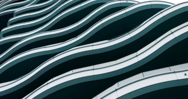 A modern building facade features undulating white balcony edges that create a flowing wave pattern across multiple floors. The dark recesses between balconies emphasize the rhythmic curves and minimalist design.