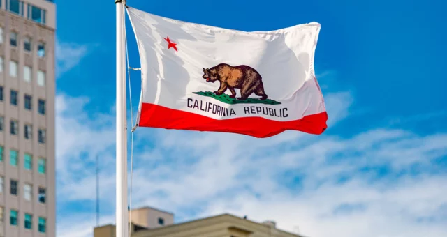 A California state flag waves against a bright blue sky, showing a brown grizzly bear walking on green grass above the words “California Republic” with a red star in the top left corner. Tall city buildings are visible in the blurred background.