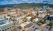 Aerial view of downtown Asheville, North Carolina, with a mix of historic brick buildings and modern mid-rises. The city is surrounded by green hills and mountains under a partly cloudy sky.