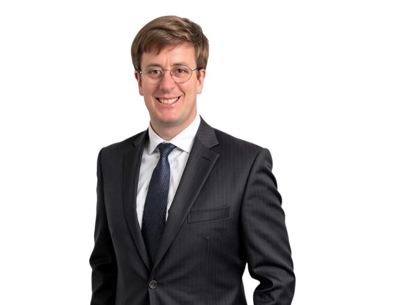 A young man in a dark pinstripe suit and navy tie stands smiling confidently. He has short brown hair, wears glasses, and is posed against a transparent background, suggesting a professional headshot or business profile photo.