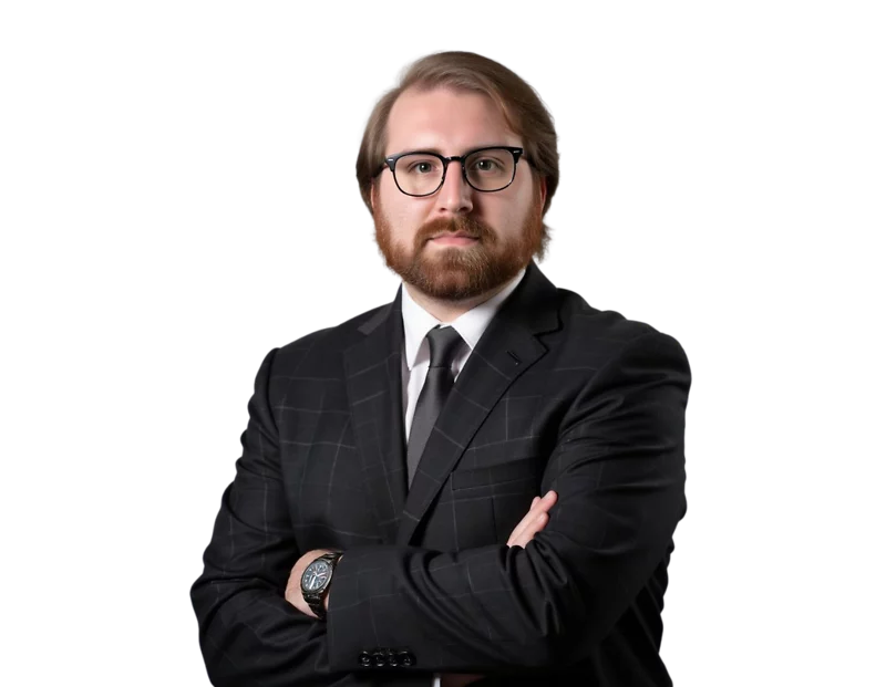 Man with a serious expression wearing a dark plaid suit, black tie, and glasses, standing with arms crossed against a transparent background. His light brown hair is neatly combed back and he has a full beard.