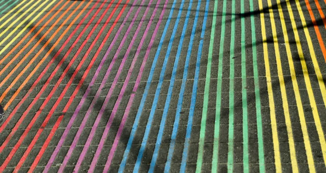 A brick-paved street is painted with evenly spaced parallel lines in rainbow colors, transitioning from red and orange to purple, blue, green, and yellow. Shadows from nearby objects fall diagonally across the vibrant pattern.