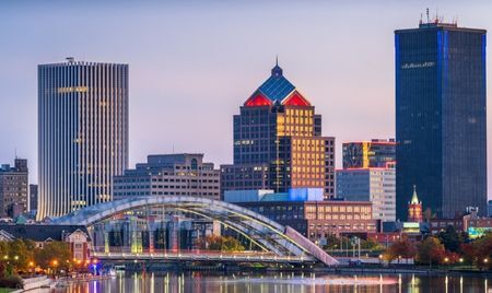 This image shows the Rochester, New York skyline at dusk, featuring tall modern buildings with colorful lights reflecting in the Genesee River. The prominent Frederick Douglass–Susan B. Anthony Memorial Bridge arcs gracefully in the foreground.