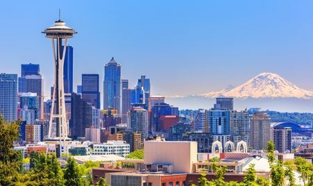 Seattle skyline on a clear day, featuring the Space Needle in the foreground and Mount Rainier in the distance. Modern high-rise buildings fill the cityscape under a bright blue sky.