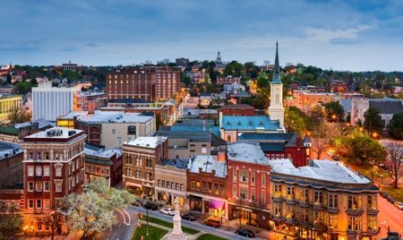 Evening view of a historic downtown with brick buildings, a church steeple, and a central square featuring a white monument. Streetlights illuminate the scene as the city stretches into the tree-covered hills beyond.