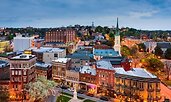 Evening view of a historic downtown with brick buildings, a church steeple, and a central square featuring a white monument. Streetlights illuminate the scene as the city stretches into the tree-covered hills beyond.