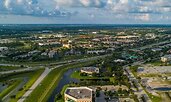 Aerial view of Port St. Lucie, Florida, showing a network of highways, commercial buildings, and residential neighborhoods. Green spaces and small waterways weave through the landscape under a partly cloudy sky.