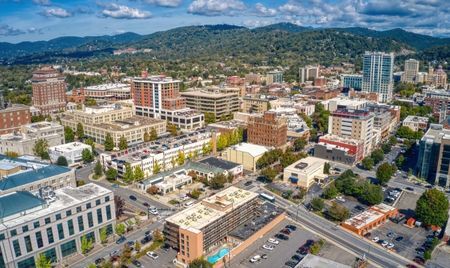 Aerial view of downtown Asheville, North Carolina, with a mix of historic brick buildings and modern mid-rises. The city is surrounded by green hills and mountains under a partly cloudy sky.