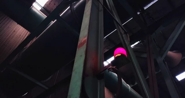 Inside an industrial facility, a bright red warning light is mounted on a metal beam among large pipes and steel framework. Dim lighting and shadows emphasize the machinery and high ceilings of the space.
