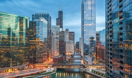 Evening view of downtown Chicago, Illinois, with illuminated skyscrapers reflecting in the Chicago River. Multiple bridges span the waterway, while the Willis Tower rises prominently in the background.