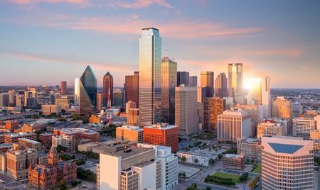 Aerial view of downtown Dallas, Texas, at sunset with sunlight reflecting off its tallest skyscrapers. The skyline features a mix of modern glass towers and historic brick buildings.
