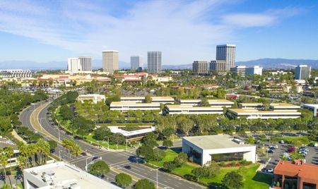 Aerial view of a business district with mid-rise office buildings surrounded by landscaped greenery and wide roads. Taller skyscrapers stand in the background beneath a bright blue sky with wispy clouds.