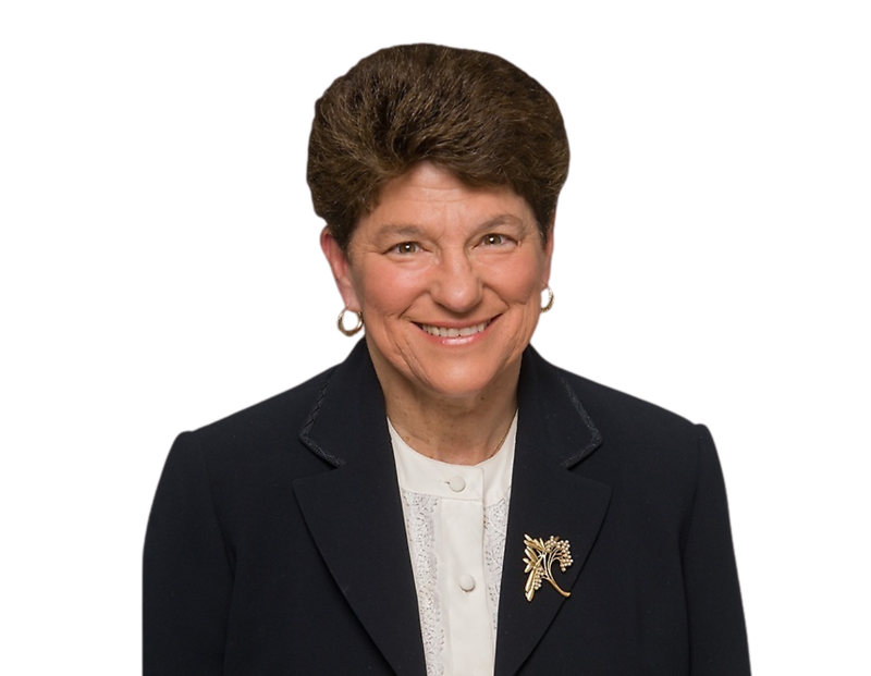 This image shows Ellen Kearns smiling warmly at the camera. She is wearing a navy blazer over a white blouse with lace detail and a decorative gold brooch shaped like a flower or branch. The background has been remove