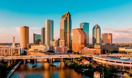 Downtown Tampa, Florida, at golden hour with sunlight reflecting off its tall glass and brick skyscrapers. A bridge crosses the Hillsborough River in the foreground, connecting the urban core with surrounding neighborhoods.