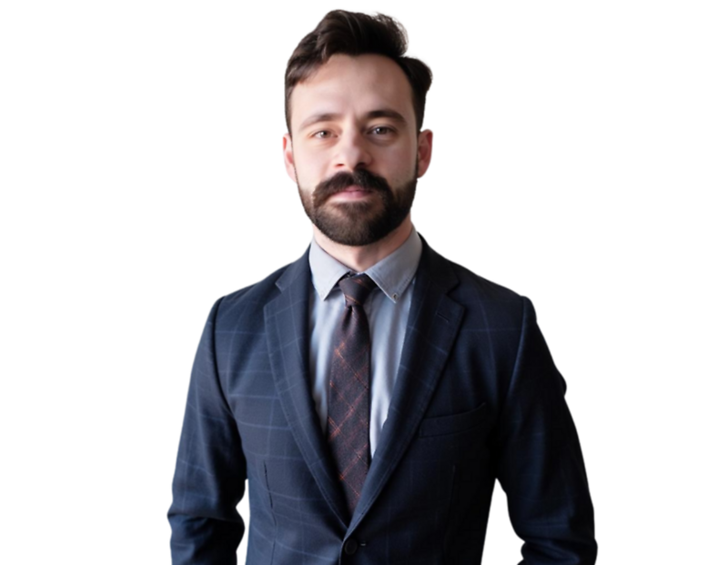 Man with short brown hair and a beard wearing a dark blue checkered suit, light blue dress shirt, and patterned tie, standing against a black background.