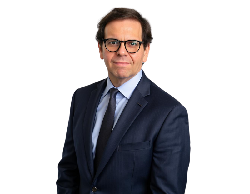 A man in a dark navy business suit with a light blue shirt and black tie poses for a professional headshot. He has short brown hair, wears black-framed glasses, and looks directly at the camera with a neutral expression.