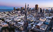 Aerial view of a city at dusk with the iconic Transamerica Pyramid rising among skyscrapers in the downtown skyline. In the foreground, tightly packed low-rise buildings lead toward the waterfront and a distant bridge.