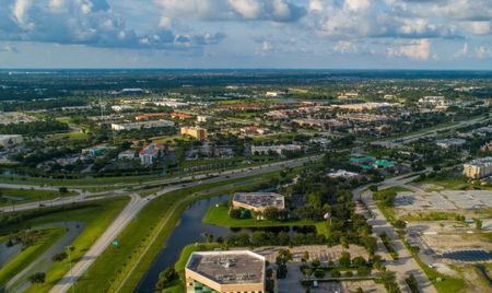 Aerial view of Port St. Lucie, Florida, showing a network of highways, commercial buildings, and residential neighborhoods. Green spaces and small waterways weave through the landscape under a partly cloudy sky.