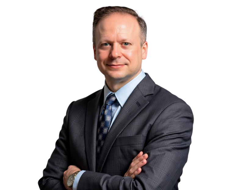Keya Denner wearing a dark plaid suit jacket, light blue dress shirt, and patterned tie, arms crossed, posed against a light blue and white geometric background.