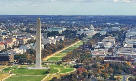 Aerial view of the National Mall in Washington, D.C., with the Washington Monument rising prominently in the foreground. The U.S. Capitol building is visible in the distance, surrounded by tree-lined streets and historic architecture.