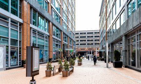 Pedestrian walkway between modern glass-and-brick buildings in the District Wharf area of Washington, D.C. The space features benches, potted plants, and a freestanding directory sign.