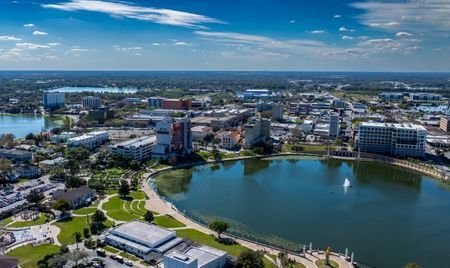 Aerial view of downtown Lakeland, Florida, featuring Lake Mirror with its central fountain. Surrounding the lake are modern buildings, green parks, and walkways under a bright blue sky.