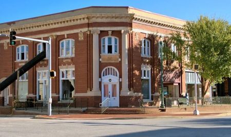 Historic red-brick corner building with white trim and arched windows, set along a quiet intersection under a clear blue sky. A tree partially shades the sidewalk beside the structure.