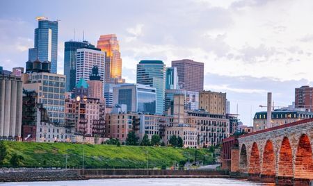 Minneapolis skyline with modern skyscrapers reflecting the evening light. The historic Stone Arch Bridge spans the Mississippi River in the foreground, leading toward the city.