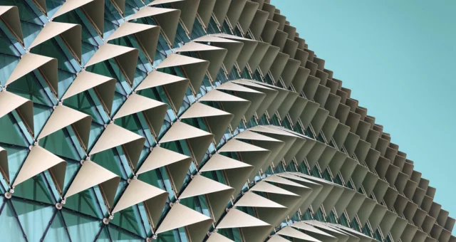 A modern building facade features rows of triangular sunshade panels angled outward from a glass exterior. The repeating geometric pattern creates a textured, scale-like effect against the clear turquoise sky.