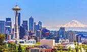 Seattle skyline on a clear day, featuring the Space Needle in the foreground and Mount Rainier in the distance. Modern high-rise buildings fill the cityscape under a bright blue sky.