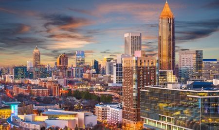 City skyline at sunset with modern high-rise buildings, including a tall tower topped with a pointed golden spire. The urban landscape is bathed in warm evening light beneath a colorful, cloud-streaked sky.