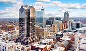 Aerial view of downtown Raleigh, North Carolina, with a mix of modern glass towers and historic brick buildings. The city stretches into the distance under a bright blue sky with scattered clouds.