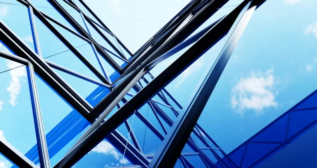 Upward view of a modern glass and steel building with angular beams reflecting the blue sky and scattered clouds. The structure's geometric lines create a dynamic, futuristic perspective.