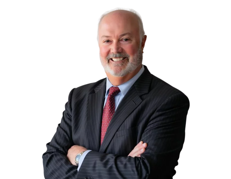 Smiling man with a gray beard and bald head wearing a dark pinstripe suit, light blue shirt, and red patterned tie. He is standing with arms crossed against a transparent background.