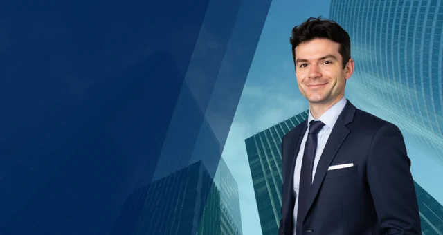 Alt text: Benjamin Rowley stands confidently in a navy suit and tie against a modern cityscape with glass skyscrapers and a gradient blue overlay. His slight smile and professional attire suggest a corporate or business-focused setting.
