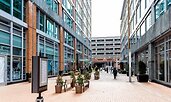 Pedestrian walkway between modern glass-and-brick buildings in the District Wharf area of Washington, D.C. The space features benches, potted plants, and a freestanding directory sign.