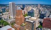 Aerial view of downtown Portland, Oregon, with a mix of modern skyscrapers and mid-rise buildings. The city is surrounded by greenery and distant hills under a clear blue sky.