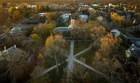 Aerial view of a college campus at sunset, with intersecting walkways lined by autumn trees. Academic buildings are spread across the landscape, bathed in warm golden light.