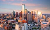 Aerial view of downtown Dallas, Texas, at sunset with sunlight reflecting off its tallest skyscrapers. The skyline features a mix of modern glass towers and historic brick buildings.