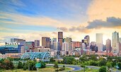 A panoramic view of Denver’s downtown skyline at sunset, with modern high-rise buildings reflecting soft golden light. In the foreground, a curved highway and green park space lead toward the city, while the blue arch of a bridge stands out against the ur