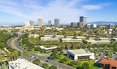 Aerial view of a business district with mid-rise office buildings surrounded by landscaped greenery and wide roads. Taller skyscrapers stand in the background beneath a bright blue sky with wispy clouds.
