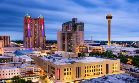 Evening view of downtown San Antonio, Texas, with the Tower of the Americas rising above the city. Colorfully lit high-rise hotels and modern buildings stand against a twilight sky.