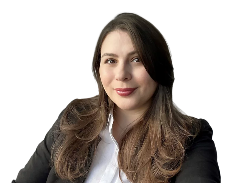 A woman with long, wavy brown hair and natural makeup, smiling softly in a professional headshot. She is wearing a black blazer over a white button-up shirt, set against a plain background.