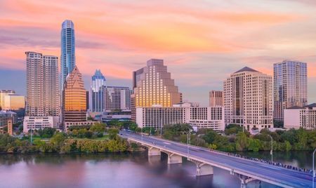 Downtown Austin, Texas, at sunset with a colorful sky casting warm light on modern skyscrapers. The Congress Avenue Bridge stretches across Lady Bird Lake in the foreground.