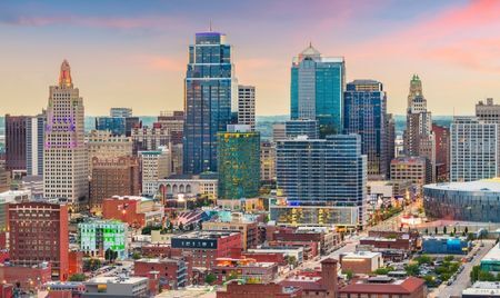 Downtown Kansas City, Missouri, at sunset with a colorful sky behind its mix of historic and modern skyscrapers. Streets below are lined with brick buildings, shops, and entertainment venues.