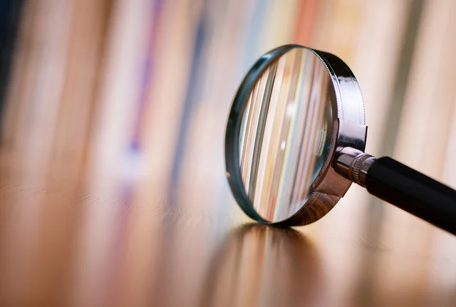 Close-up of a magnifying glass lying on a wooden surface, focusing on the blurred vertical lines of books in the background. The lens distorts and enlarges the colorful spines, symbolizing investigation, research, or attention to detail.